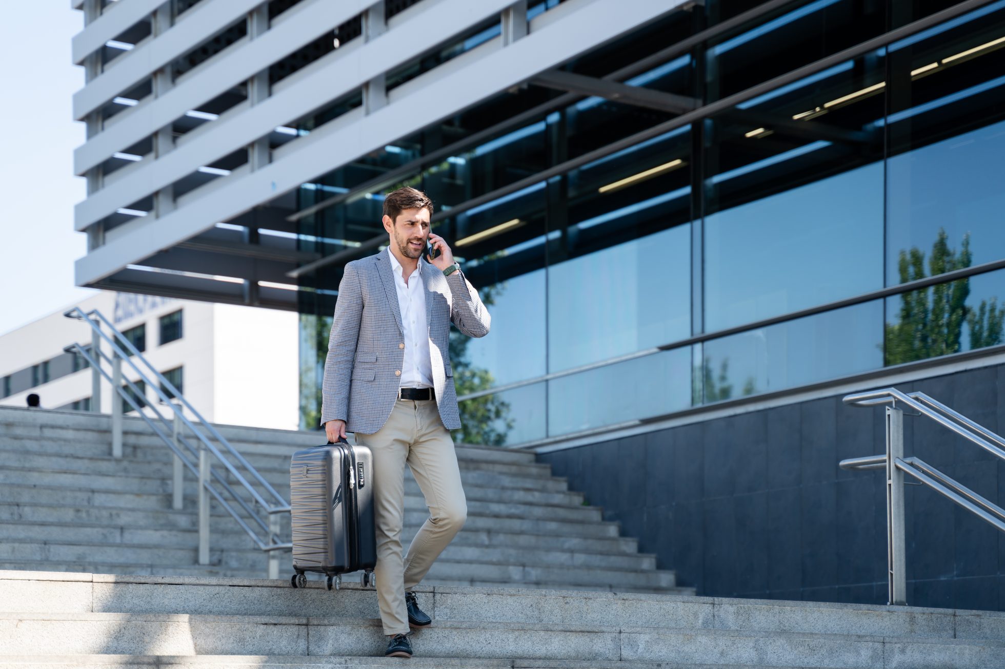 A businessman walks down the stairs of a modern office building, talking on the phone and dragging a suitcase behind him.